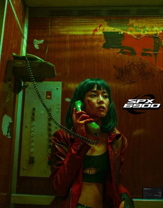 A woman with green hair and a red jacket holds a phone receiver in a graffiti-covered elevator, with 'SPX 6900' branding visible.