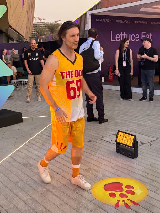 A person wearing a yellow and orange basketball jersey with 'THE DOG 69' printed on it and matching shorts, posing at what appears to be a tech event with 'BREAKPOINT' and 'Lettuce Eat' signage visible in the background.