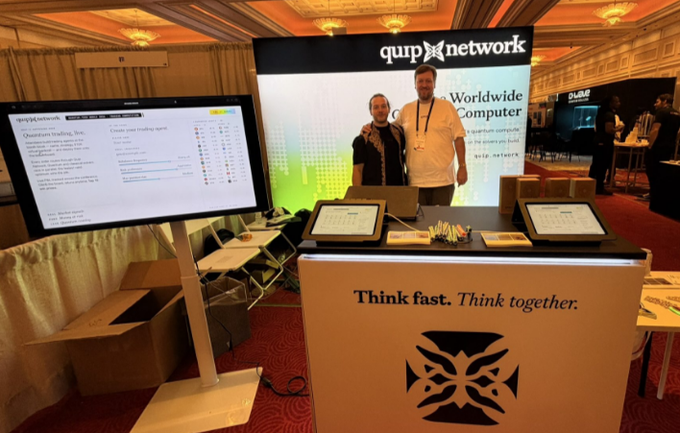 Two people stand smiling at the Quip Network booth, which displays a large screen with the "quip network" logo and "Worldwide Computer" text, alongside a counter featuring iPads and the tagline "Think fast. Think together." The setup reflects the post's reference to Quip Network, where visitors created their own trading problems and competed in 10-second portfolio races with 1,930 live contests running in the background.