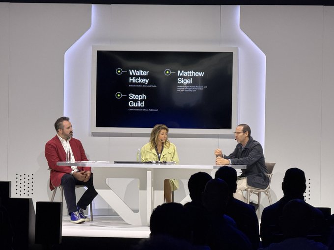 A panel discussion with three people seated at a white table on a stage, with a screen behind them listing speakers Walter Hickey, Matthew Sigel, and Steph Guild.