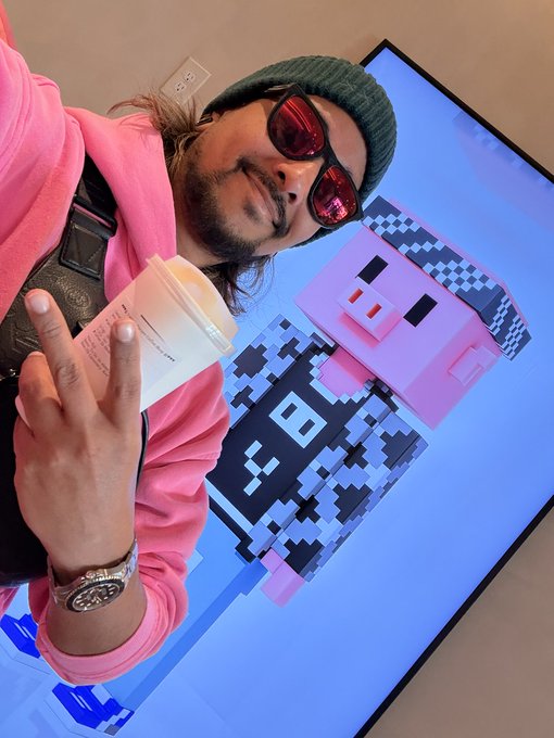 A person wearing a green beanie, red sunglasses, and a pink hoodie holds up a peace sign while standing in front of a large display featuring a pixel-art pink axolotl character.