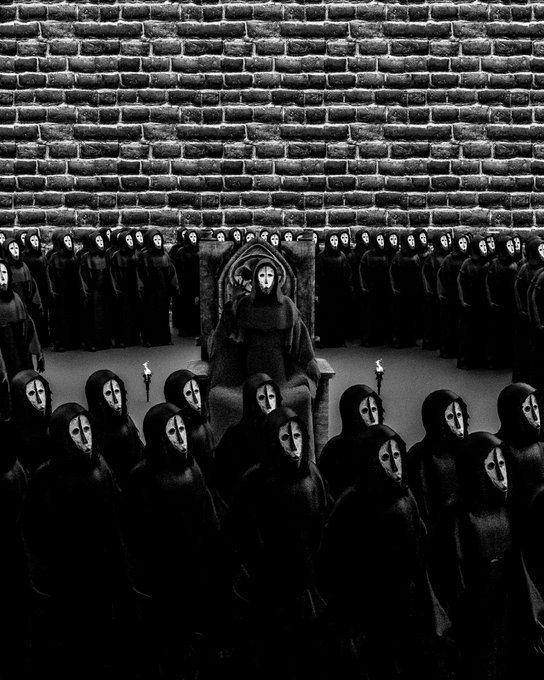 A black and white, eerie illustration of a crowd of hooded figures wearing white masks, with one central masked figure sitting on an ornate throne against a brick wall background.