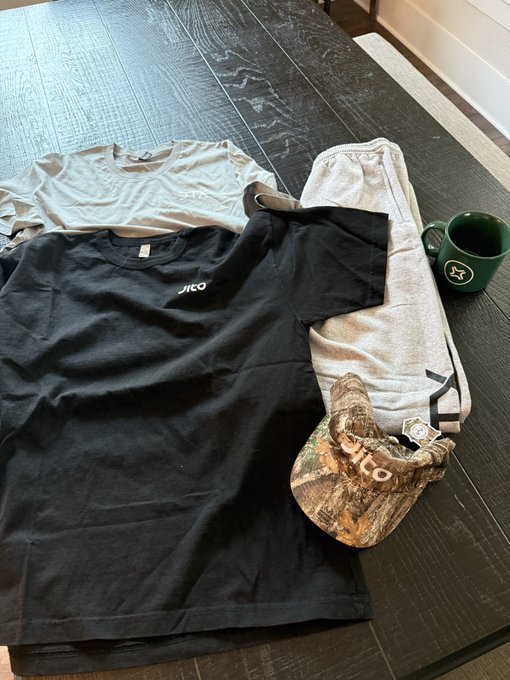 A casual outfit laid out on a dark wood floor, consisting of a white t-shirt, a black t-shirt with 'NITCO' text, gray sweatpants, a camouflage hat, and a green ceramic mug.