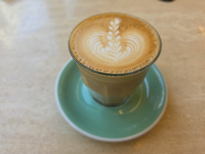 The image features a cup of coffee with latte art on top sitting on a saucer, placed on a table. The coffee appears to be freshly made and has a beautiful design created by the latte art. The background of the image is blurred, drawing attention to the cup of coffee as the main focus.