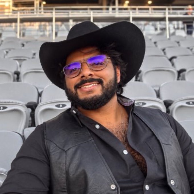 A selfie of a smiling man wearing a black cowboy hat, purple-tinted sunglasses, and a black leather vest, taken in an empty stadium seating area.