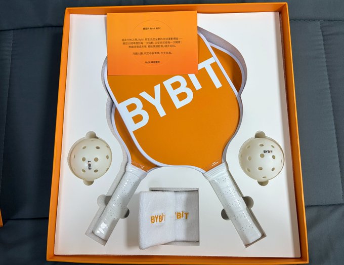 An orange gift box set from Bybit featuring two paddle-shaped items and two balls branded with the Bybit logo.