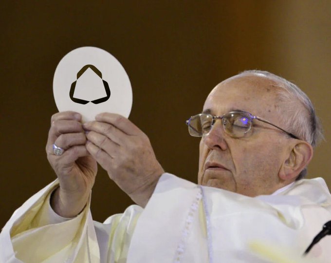 Pope Francis holds up a white communion wafer featuring a triangular recycling logo, symbolizing a consecration. The image's juxtaposition of sacred ritual with a corporate sustainability symbol creates a striking visual metaphor, though it has no direct connection to the post's discussion of the $TIG ticker and its underappreciated risk/reward potential in the crypto market.