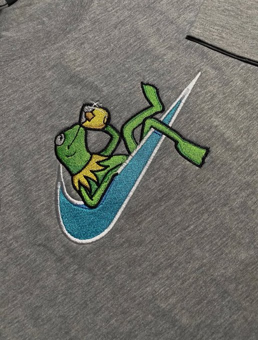 A Nike logo embroidered on gray fabric, featuring the iconic green frog character Kermit the Frog lounging on the teal swoosh.