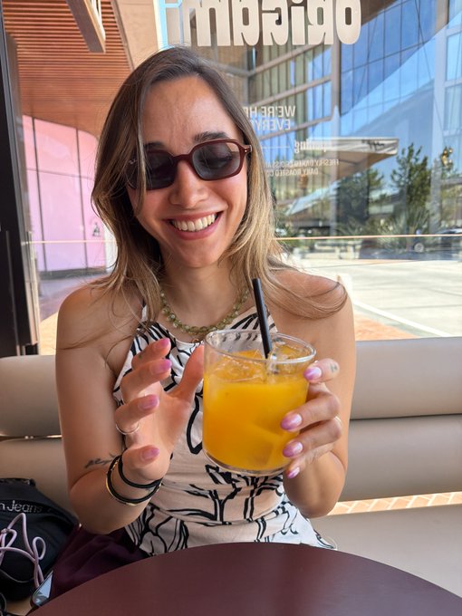 A smiling woman with sunglasses holding up a glass of orange juice, taken from across a table inside what appears to be a restaurant or cafe.