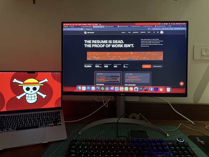 A desk setup featuring a MacBook with a Straw Hat Pirates logo wallpaper next to a monitor displaying 'THE RESUME IS DEAD. THE PROOF OF WORK ISN'T.' with Proof of Work stats including 16,984 and 190, above a backlit RGB gaming keyboard.
