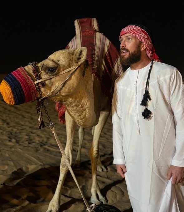 A man in traditional white Emirati attire with a red-checkered keffiyeh stands beside a decorated camel in a moonlit desert, projecting confidence and vision. The image embodies the post's bullish optimism—signaling a "gm" (good morning) with the steady, forward-looking conviction of someone who sees only good things ahead on the horizon.