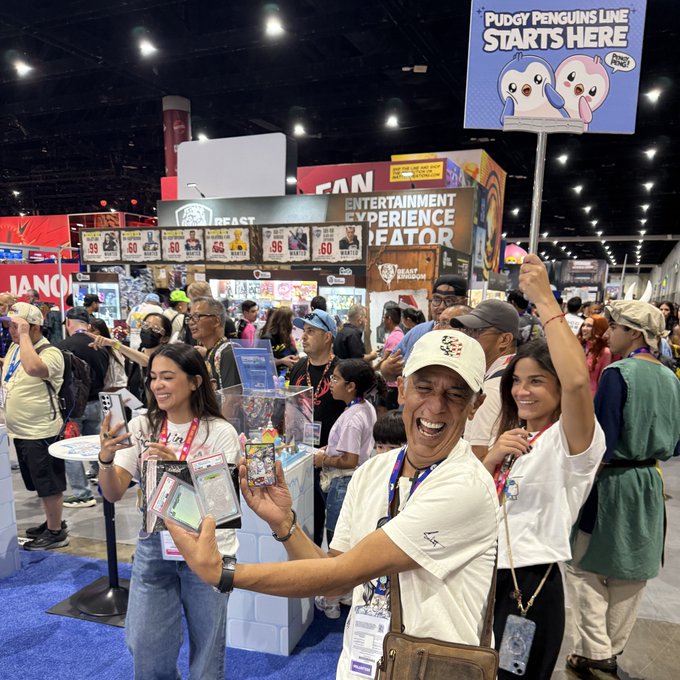 Three people at a convention smile and hold up Pokémon merchandise, with a sign above reading "PUDGY PENGUINS LINE STARTS HERE."