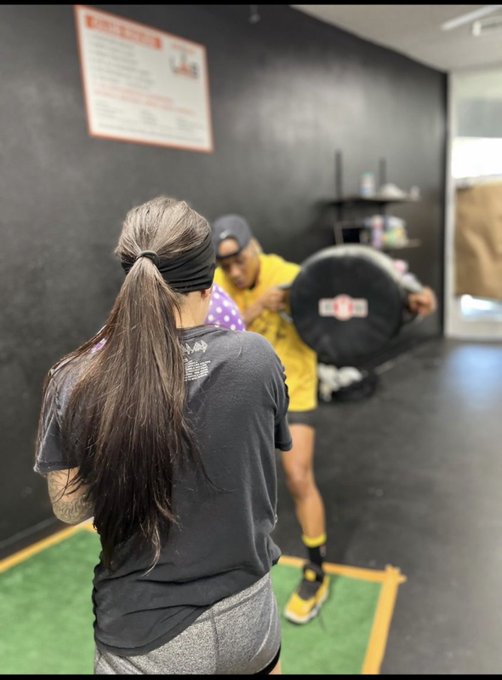 A woman with a long ponytail and black headband watches as a trainer in a yellow shirt holds a circular "TITLE" branded punching pad during a boxing training session in a gym with artificial turf. The image captures the practical demonstration referenced in the caption "This is how I bagwork," showing the bagwork technique in action rather than using a traditional heavy bag.