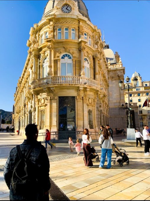 A grand, ornate yellow stone building with a domed clock tower stands prominently in a sunny plaza, where people are walking, including a person in the foreground wearing a black jacket and backpack.