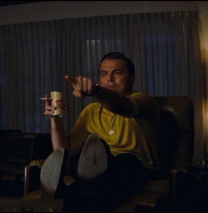 A man in a yellow shirt sits in a chair holding a beer can while pointing at the camera, with his feet propped up.