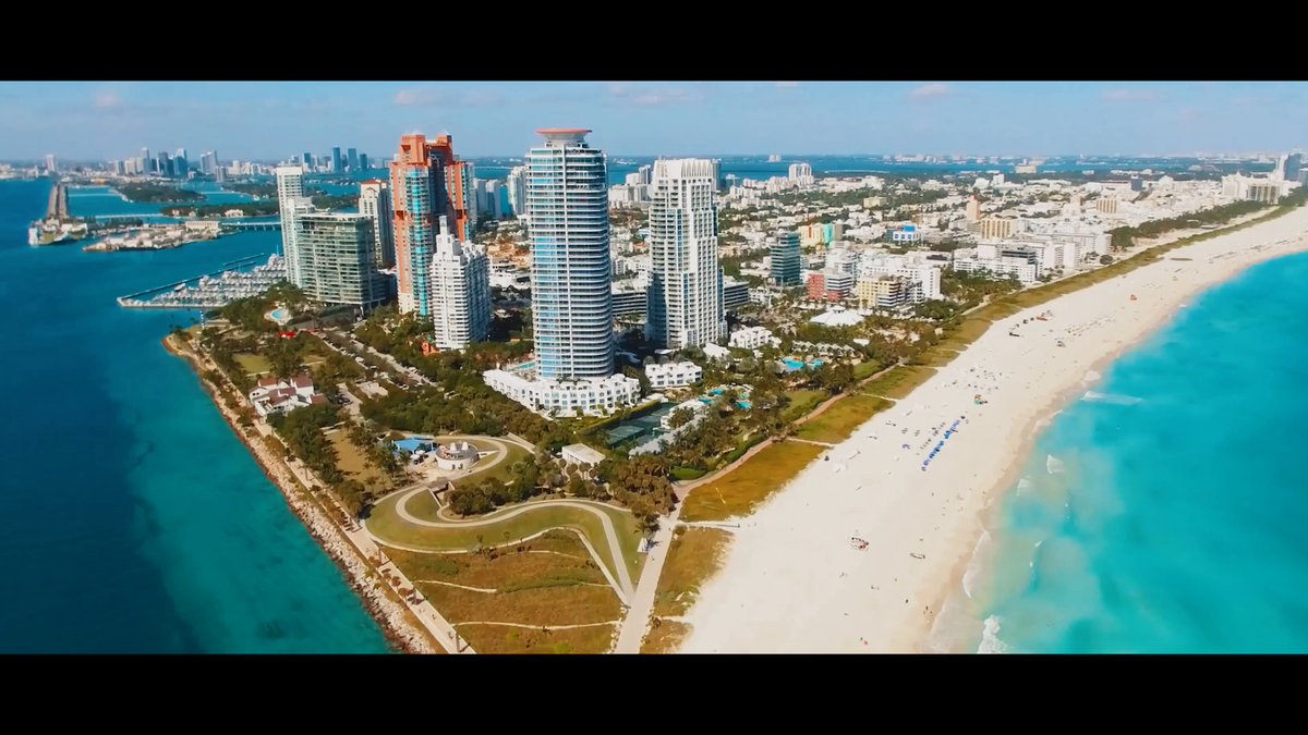 The image shows an aerial view of a city with tall buildings along the shoreline. There are several people visible near the water's edge, enjoying their time at the beach. A boat can be seen floating nearby. The sky above appears blue with some clouds scattered across it. The overall scene conveys a sense of relaxation and leisure as people spend their day by the ocean.