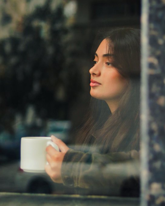 A young woman with long dark hair in profile, looking out a window while holding a white mug.