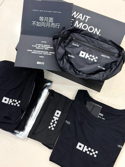 A flat-lay display of OKX-branded Mid-Autumn Festival gifts, including a black fanny pack marked "24/7/365," folded black athletic wear with the OKX logo, and a card reading "WAIT FOR THE MOON" alongside the Chinese poem "等月圆 不如向月而行" ("Instead of waiting for the moon to be full, why not walk toward the moon"). The sleek black apparel and accessories reflect the post's motivational message about taking action toward goals rather than waiting for the perfect moment.