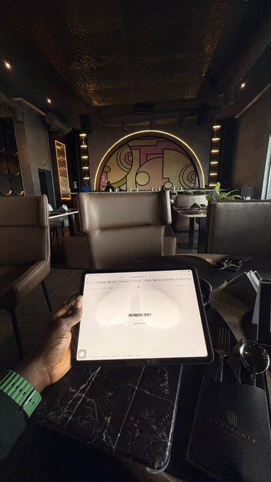 A first-person view inside a dimly lit lounge or restaurant shows a hand holding a tablet displaying a menu with 'MEMBERS ONLY', with a dark marble table, brown leather seating, and an arched decorative wall in the background.