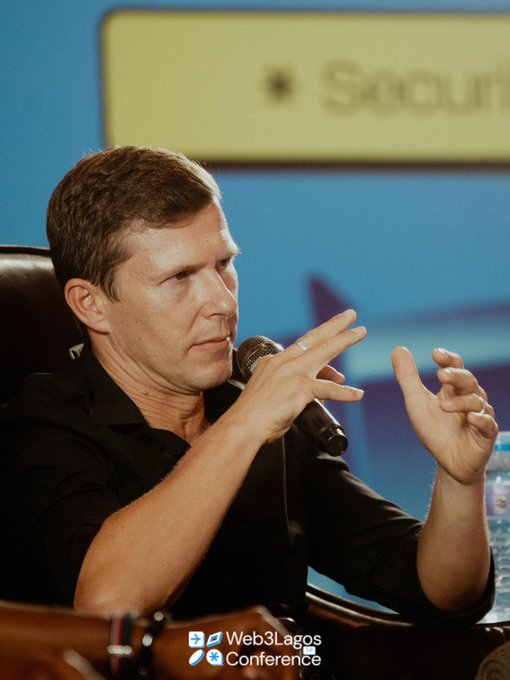A man in a black shirt speaks into a microphone at the Web3Lagos Conference, gesturing with his hands while seated against a blue background.