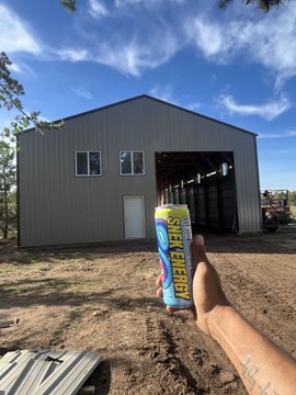 The image shows a person holding up a can of soda, which has the word "Snek" written on it. The background features an unfinished building with a gray roof and windows.