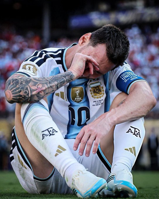 Lionel Messi (#10, Argentina) sits on the pitch in his World Cup jersey, wiping his face with his tattooed arm.