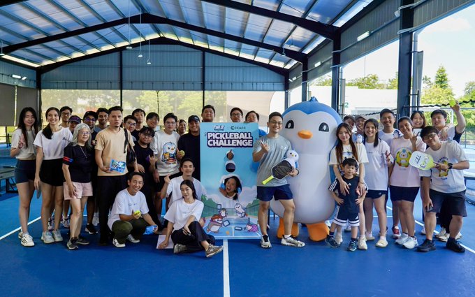 A large group photo of people posing at a pickleball event, with a banner reading 'PICKLEBALL CHALLENGE' and a large penguin mascot standing among the participants.