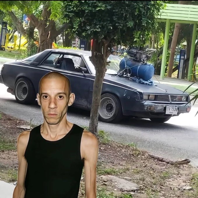 A bald, thin man in a black tank top stands in the foreground in front of a gray vintage car with a blue gas tank mounted on the roof, parked on a street near a park.