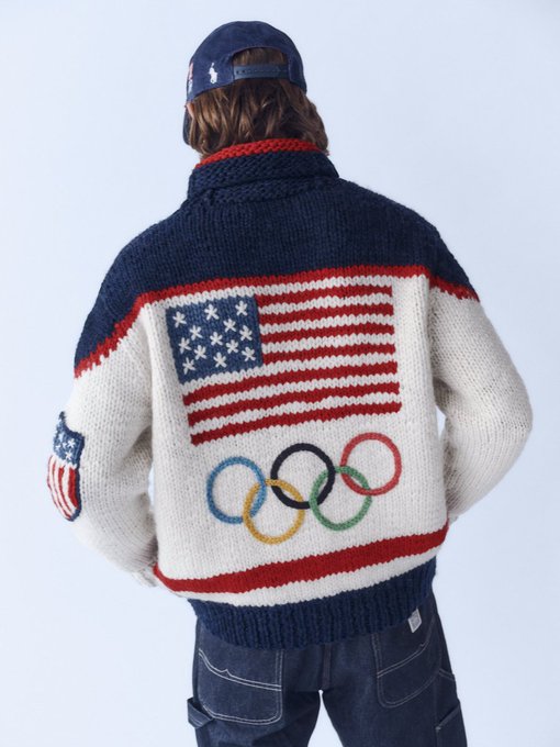 A rear view of a person wearing a navy blue and cream knit cardigan featuring a large American flag design on the back and the Olympic rings below it, paired with a baseball cap and jeans.