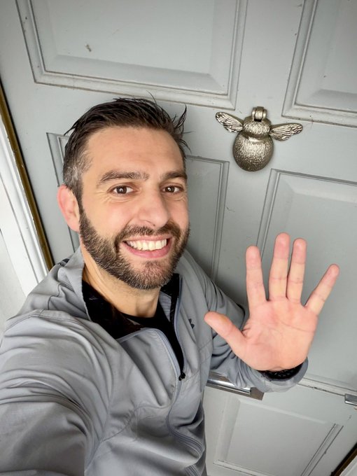 A selfie of a smiling bearded man in a gray jacket holding up his hand in front of a gray door with a decorative bee door knocker.