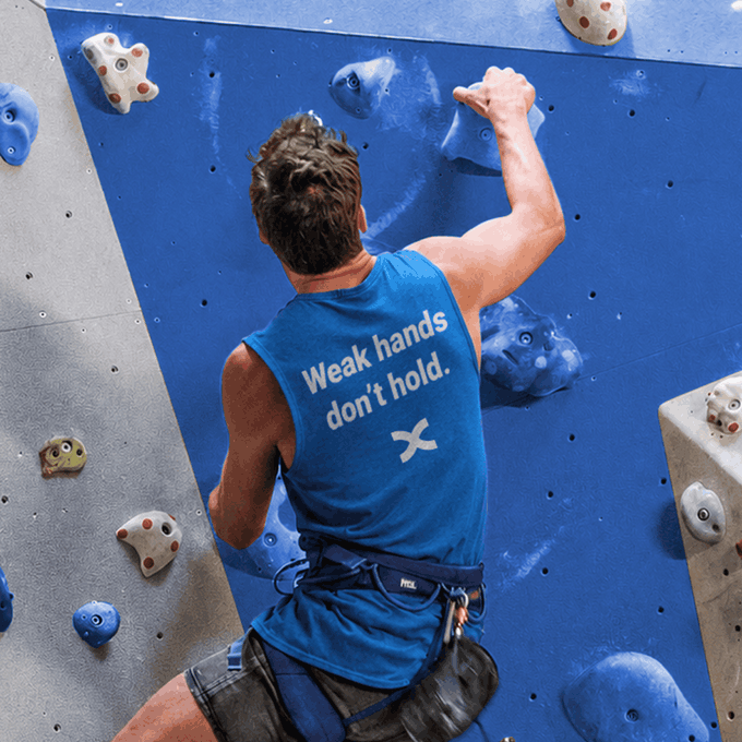 A rock climber grips blue and gray climbing holds on an indoor wall, wearing a blue tank top that reads "Weak hands don't hold." The shirt's message parallels the post's reminder to stay steady and maintain your grip even after a disappointing red candle in the market.