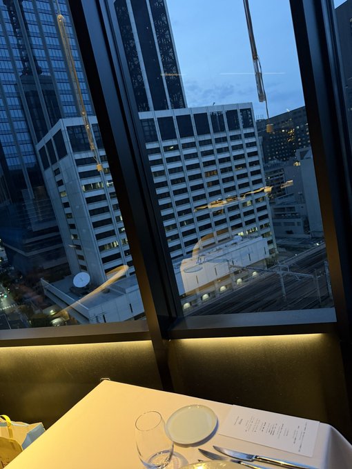 A French restaurant table with a wine glass and menu overlooks the Ginza skyline at dusk. The refined urban setting contrasts with the post's casual chatter about crypto meme drama and social media gossip.