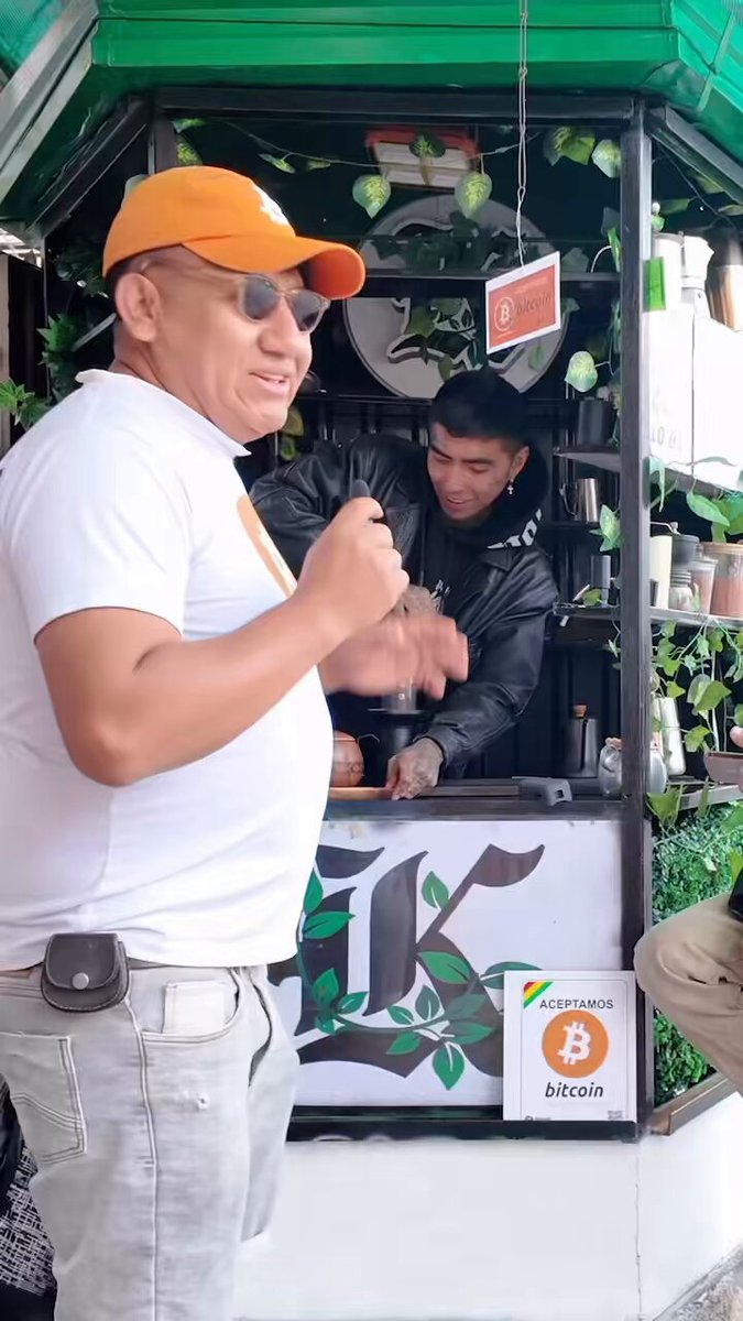 The image shows a man wearing an orange hat standing next to a green building with a sign that says "Bolivia Hoy visitamos la segunda sucursal de Bitcoin en la Plaza Uyuni." The man appears to be in front of a coffee shop, and there is another person inside the shop. A post on social media indicates that this location is a Bitcoin Cafe.