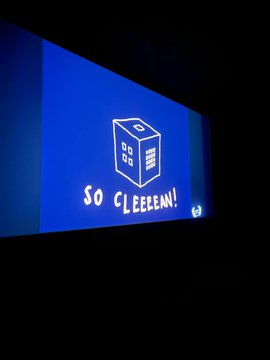 The image features a large screen displaying an advertisement or trailer for a movie called "So CLEEEN!". The screen has a blue background with white text that reads "SO CLEEEN!", accompanied by a small cube logo, possibly representing Bitcoin. The scene takes place in front of a building, and there is a person visible on the right side of the image.