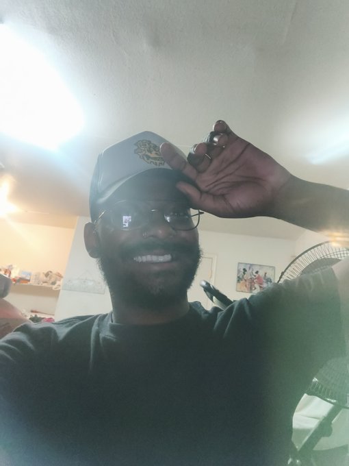 A smiling man wearing a baseball cap and glasses poses for a selfie indoors, touching the brim of his hat with his hand.