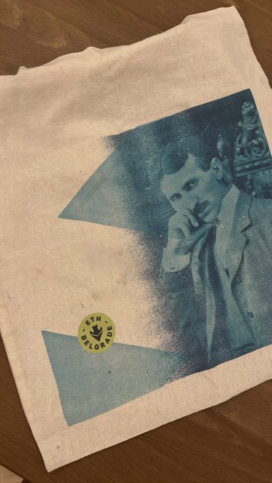 A white t-shirt laid on a wooden surface with a blue-toned print of a young man in a suit with his hand on his chin, accompanied by a small yellow 'ETH VELORACE' sticker, which appears to reference Ethereum founder Vitalik Buterin.