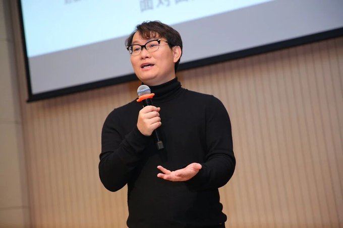 An East Asian man wearing glasses and a black turtleneck speaks into a microphone while gesturing with his hand, standing in front of a projection screen.