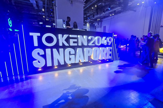 A large illuminated sign reads "TOKEN2049 SINGAPORE" at a crypto conference event, with attendees gathered in the background bathed in blue lighting.