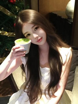 A photograph of a young woman with long dark hair wearing a white dress, holding a cup of matcha latte, posed in front of a Christmas tree.