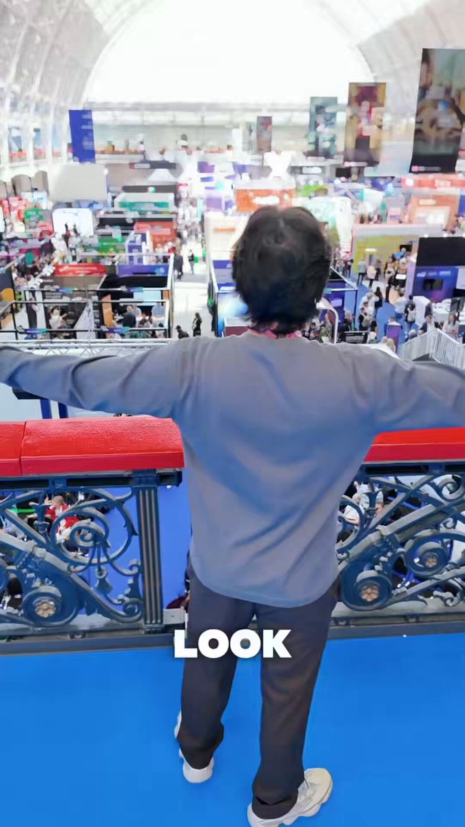 The image shows a person standing on a balcony, looking out over a large convention center filled with booths and displays. They are wearing a gray sweatshirt and pants while holding their arms wide open. The caption of the image reads "I'm at Solana Breakpoint London early! Well not exactly... come with me through a tour of The Olympia The venue hosting the hottest crypto event later this year". This suggests that the person is attending or has attended an event related to cryptocurrency, possibly exploring the convention center and learning more about it.