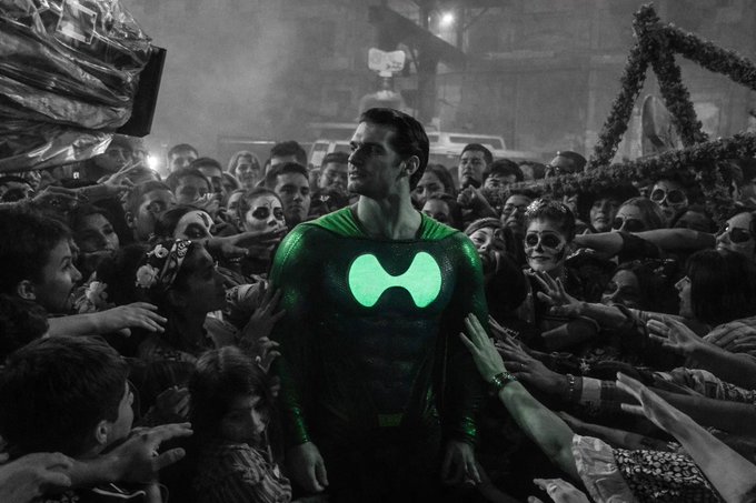 A man in a green superhero costume with a glowing 'H' logo stands in a black-and-white festival crowd wearing Day of the Dead face paint.