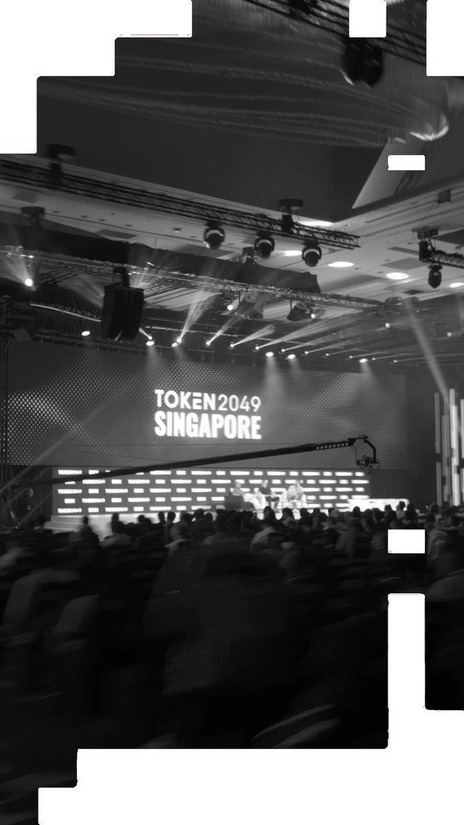 A black-and-white photo from the TOKEN2049 Singapore conference shows a large auditorium filled with attendees seated before a brightly lit stage displaying the "TOKEN2049 SINGAPORE" logo, with professional lighting rigs and a camera crane suspended overhead. The image captures the scale and atmosphere of the event referenced in the post about sharing a vision for convergence across crypto, AI, and TradFi.