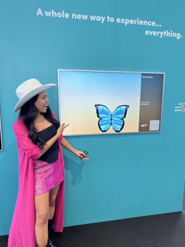 A woman wearing a white hat and a bright pink coverup points at a butterfly graphic displayed on a screen, with the text 'A whole new way to experience... everything.' visible on the teal wall behind her.