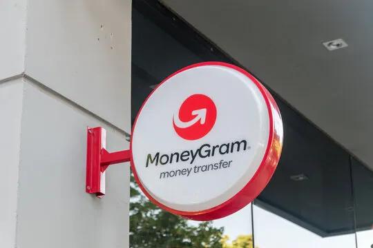 The image features a red sign with white text that says "MoneyGram" above a logo of a red circle with an arrow pointing upwards, which is part of the MoneyGram stablecoin. The sign is attached to a building or wall using a bracket and is positioned on the side of the structure.
