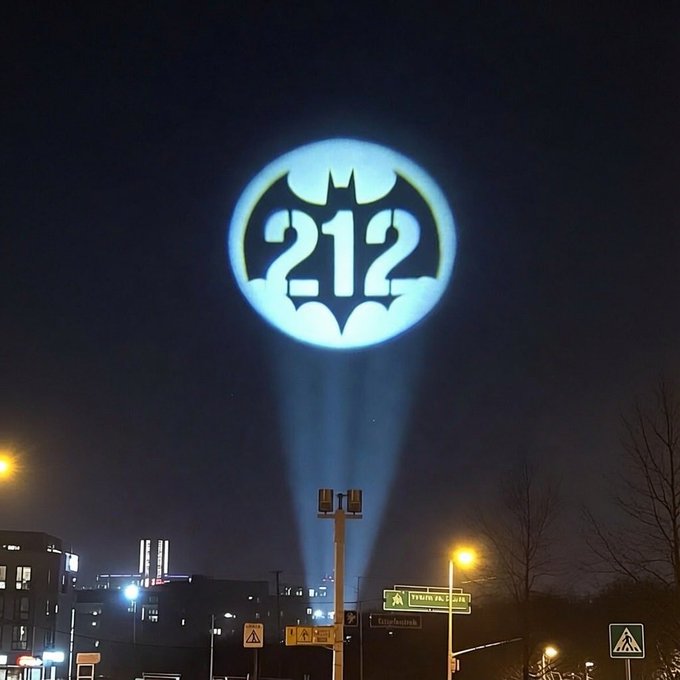 A nighttime photograph shows the Batman signal projected into the sky, but with the number '212' incorporated into the bat logo instead of the usual shape, shining over a city street.