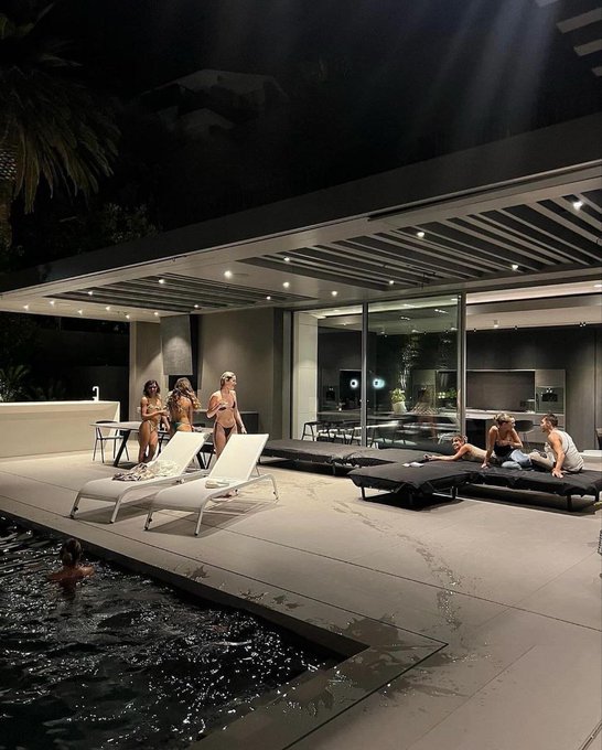 A nighttime poolside scene at a modern luxury home where several women in swimwear gather near lounge chairs and daybeds by the illuminated swimming pool.