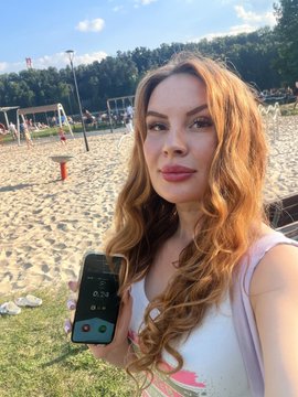 A woman with long reddish-blonde hair takes a selfie on a sandy beach while holding up a smartphone displaying a running timer app.