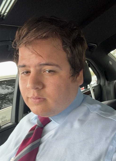A selfie of a young man with brown hair wearing a light blue dress shirt and a red and blue striped tie, sitting in the driver's seat of a car.