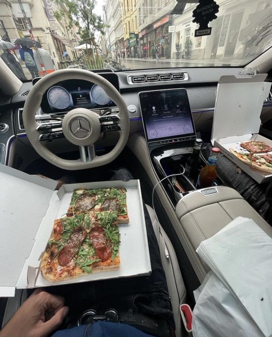 Interior view of a Mercedes-Benz car showing a pizza box with a sliced pizza topped with arugula and pepperoni resting on the driver's lap, with a rainy street scene visible through the windshield.