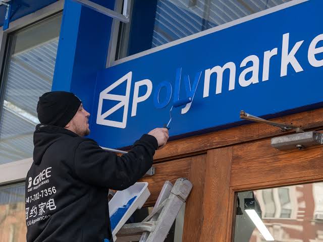 The image shows a man painting a blue sign for Polymarket outside of a building, using a roller brush. He appears to be working diligently to cover the entire surface of the sign with paint. The background features a window that provides some context about the location or business associated with the sign.